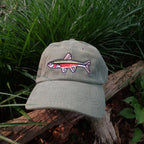 A light olive green corduroy baseball cap with a colorful embroidered fish patch on the front, resting on a fallen log surrounded by green grass and forest floor foliage.